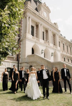 Wedding party postures casually on a grand lawn in front of an elegant white historic mansion with classical columns.