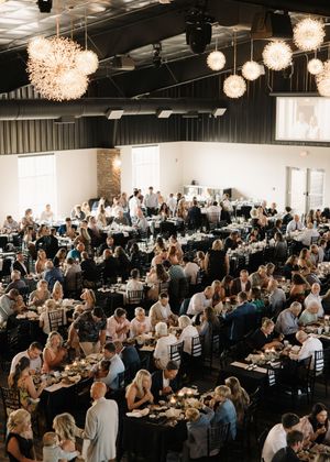 A large wedding reception hall filled with guests dining at round tables under elegant crystal chandeliers and modern lighting.