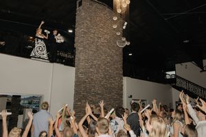 Crowd of concert-goers with raised hands below dark ceiling with dangling lights in a music venue.