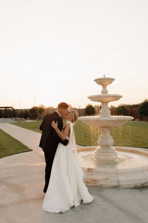 Newlywed couple shares romantic kiss by elegant tiered fountain at sunset in outdoor garden setting.