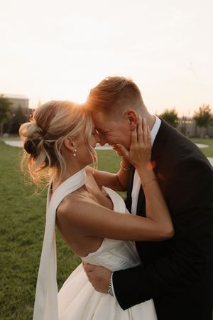 Romantic wedding photo of couple embracing during golden hour sunset in a grassy field with soft backlight.