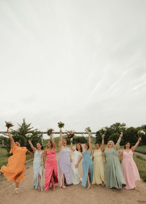 A festive bridal party wearing colorful pastel dresses poses together on a scenic outdoor pathway.