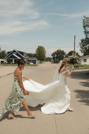 A bridesmaid in a floral dress helps adjust a flowing white wedding gown train on a sunny suburban street.