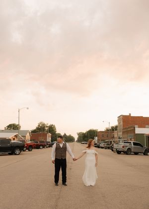 A couple holds hands while standing in the middle of a quiet small town street at sunset with vintage cars parked along both sides.