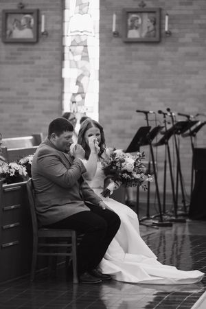 Black and white photo of emotional couple sitting together at wedding ceremony with flowers and microphone stands in background.