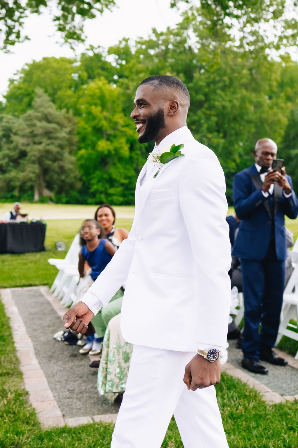 Groom in all-white suit showing joy during outdoor wedding ceremony.