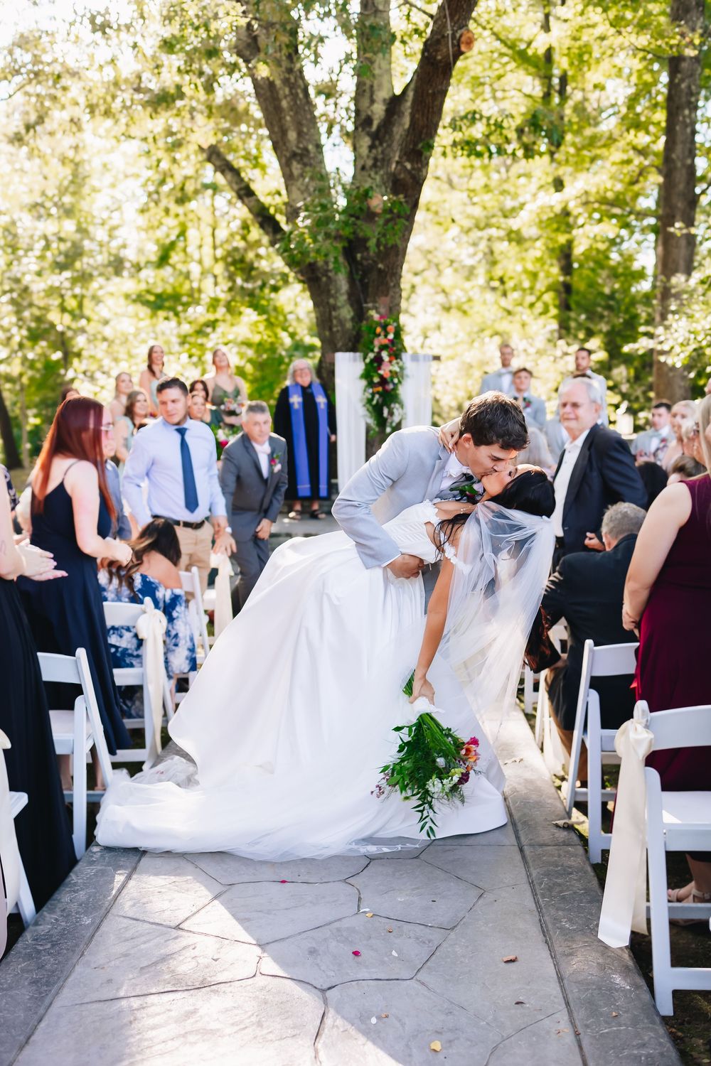 Wedding party and guests pose with flowing white veils during an outdoor ceremony under trees.