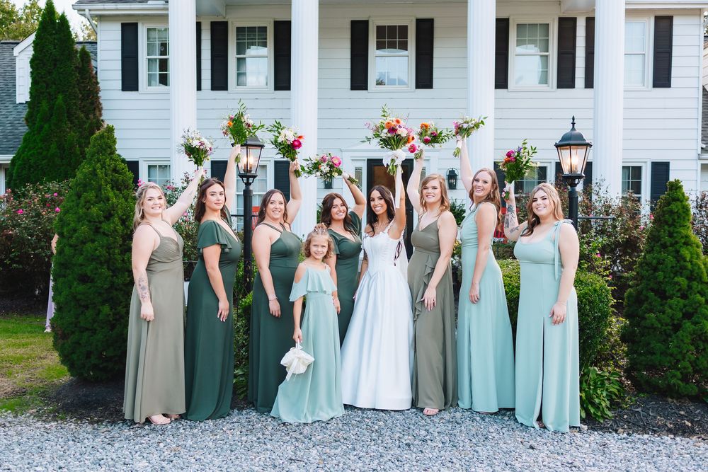 Bridesmaids in mint and beige dresses stand together against a white house backdrop.