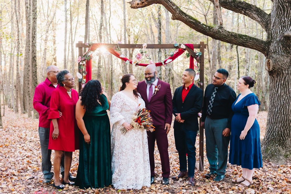 Wedding party poses under decorated arch in forest wearing deep jewel-toned formal attire.