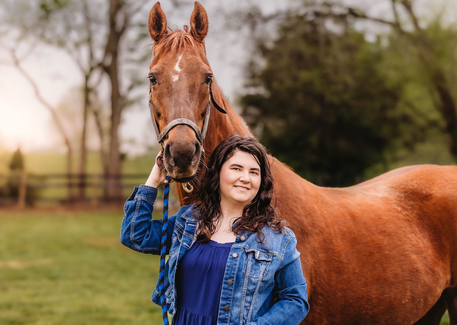 Woman in denim jacket stands with chestnut brown horse in green pasture during golden hour sunset.