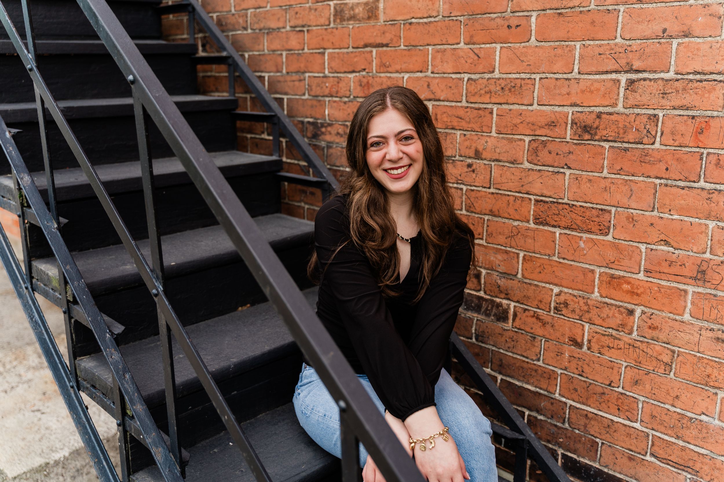Someone sits on outdoor metal stairs by a red brick wall wearing a black sweater and blue jeans.