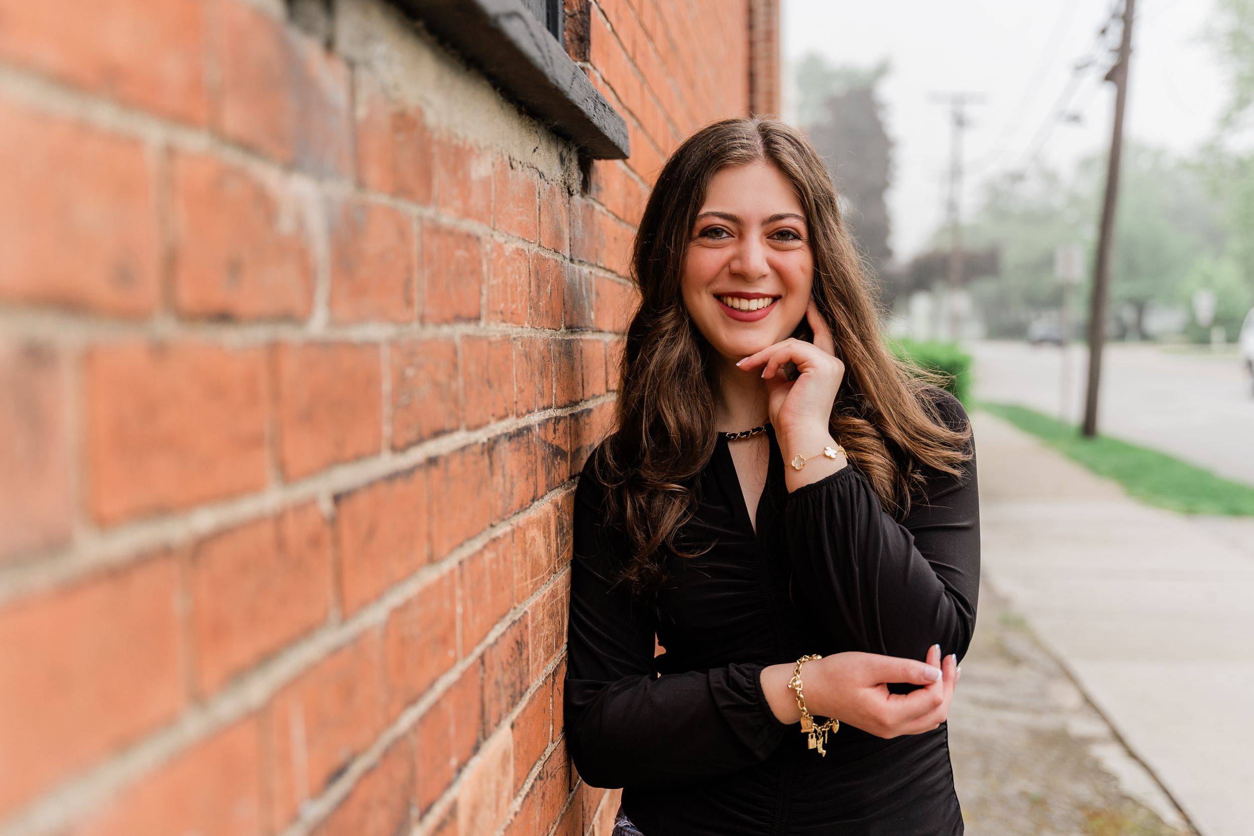 Woman in black outfit sits casually against brick wall while smiling outdoors.