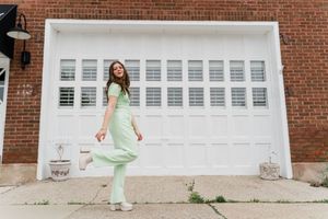 Person in mint green outfit poses playfully in front of a white garage door on brick building.