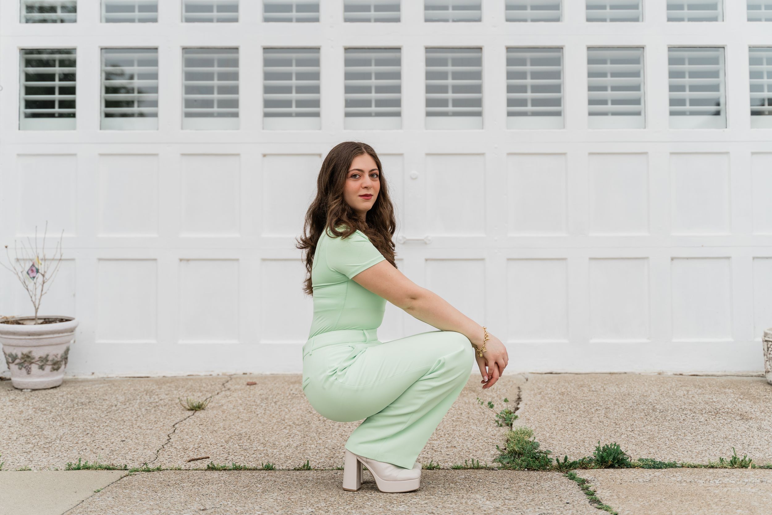 A person in a light green dress crouches on the sidewalk against a white industrial building with windows.