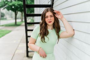 A person in a mint green dress poses against a white house exterior near stairs on a sunny day.