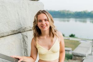 A young woman in a yellow sundress poses outdoors against concrete pillars near a waterfront setting.