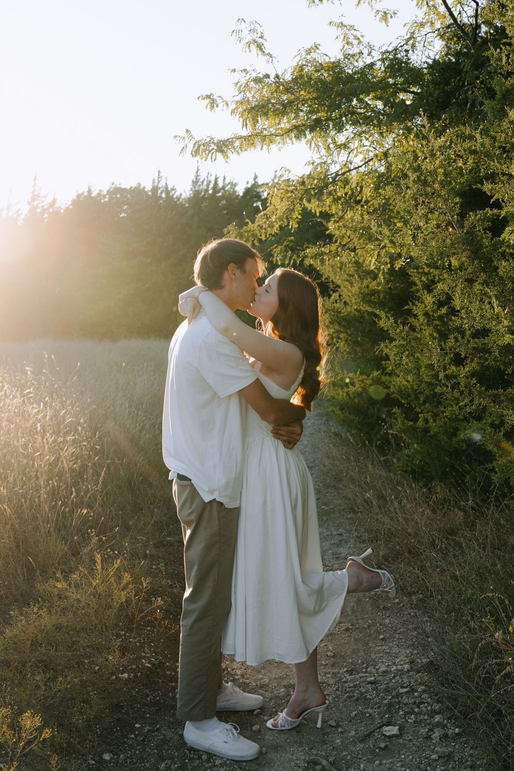 A romantic couple sharing a tender kiss at sunset in a natural outdoor setting with golden light filtering through trees.