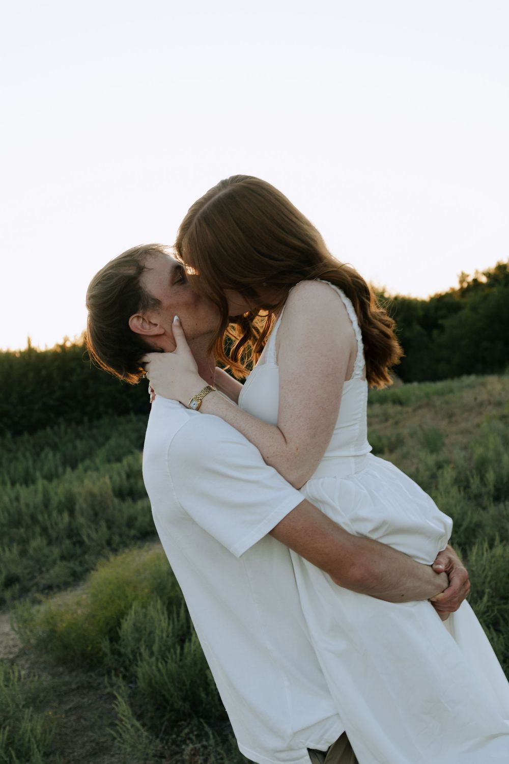 Couple in white clothing sharing passionate embrace against sunset backdrop in field.