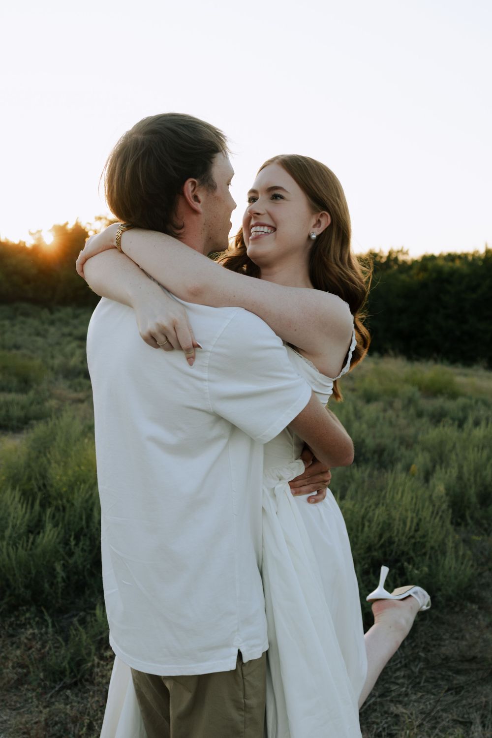 A tender embrace between a couple in white clothing against a sunset backdrop in nature.