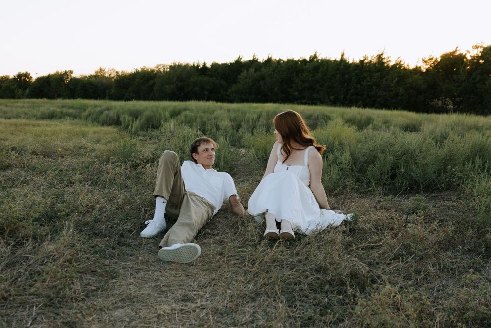 A couple in wedding attire sits together in a grassy meadow at sunset creating a romantic silhouette.