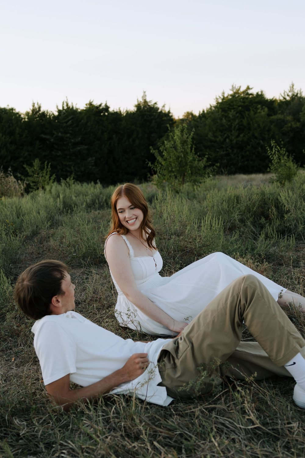 A couple lying in a grassy field at sunset share playful and intimate moments in a casual outdoor setting.