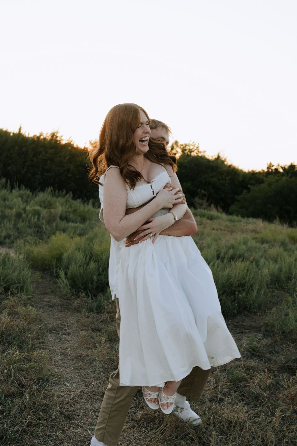 Joyful couple laughing and embracing while wearing white dresses in a rustic outdoor setting.