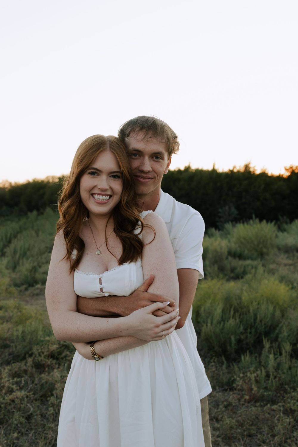 A couple embraces while posing together in a field during golden hour with greenery in the background.