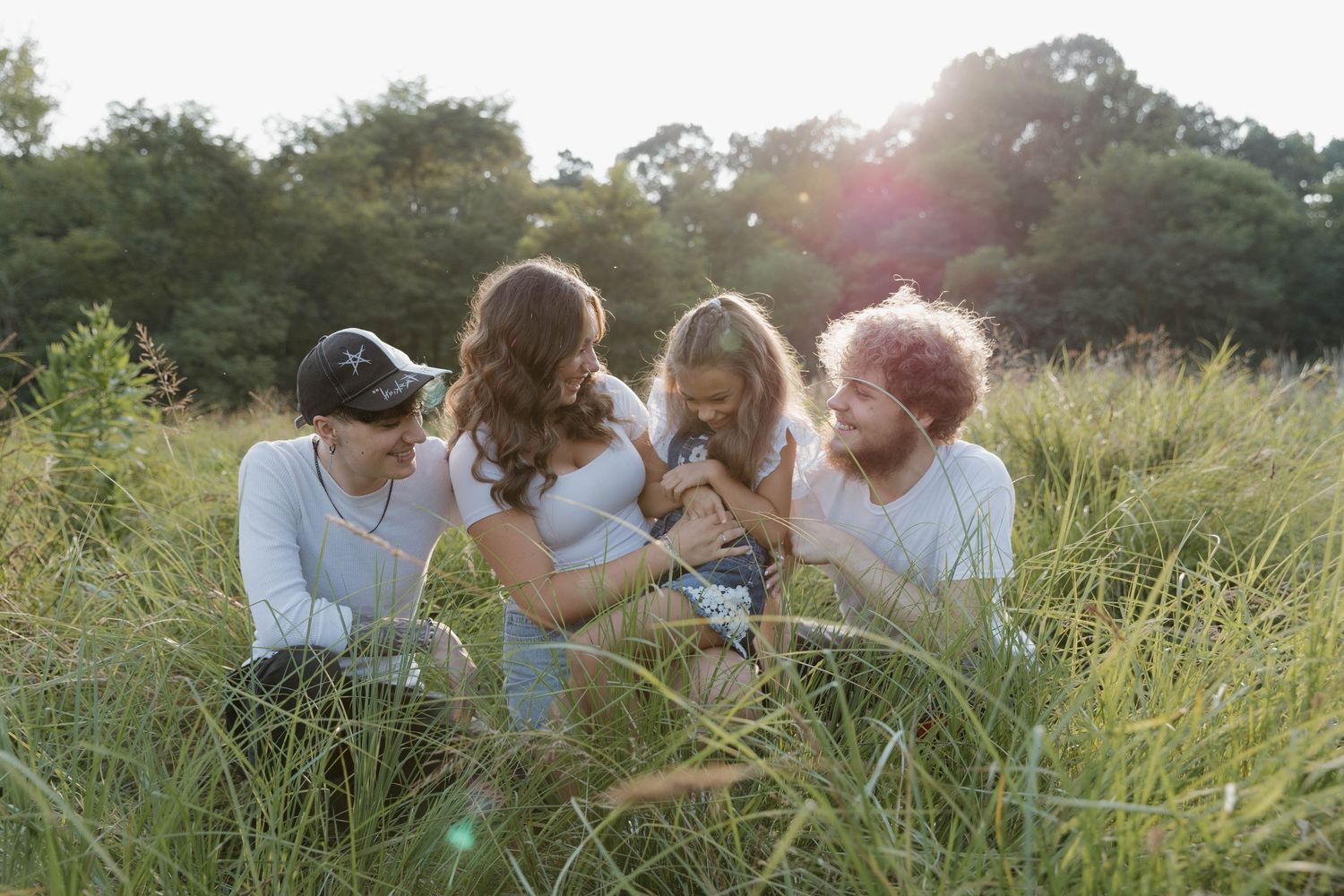 A group sits together in tall grass during golden hour as sunlight streams through trees in the background.