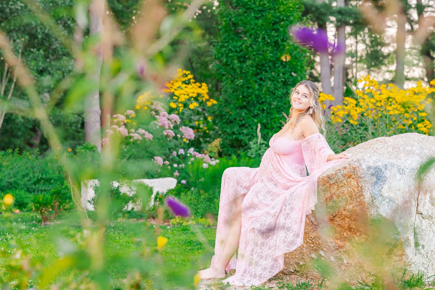 A person in a pink flowy dress sits on a rock surrounded by a vibrant garden with yellow and purple flowers.