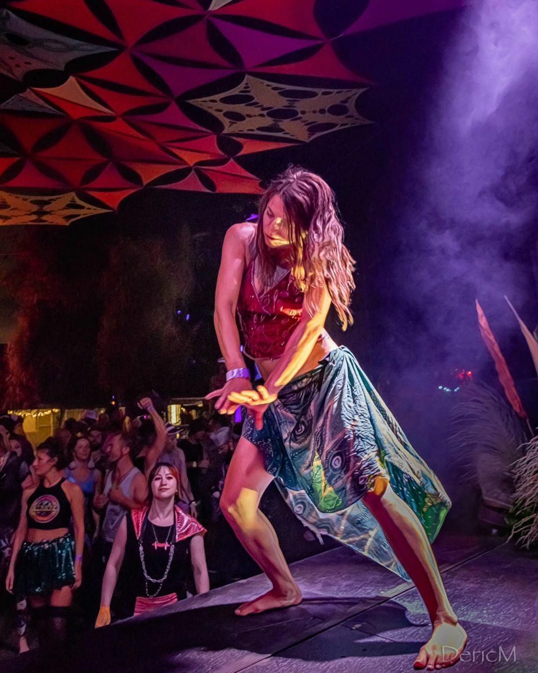 A dancer in flowing colorful clothing performs energetically at a nightclub under purple lighting and geometric ceiling decor.