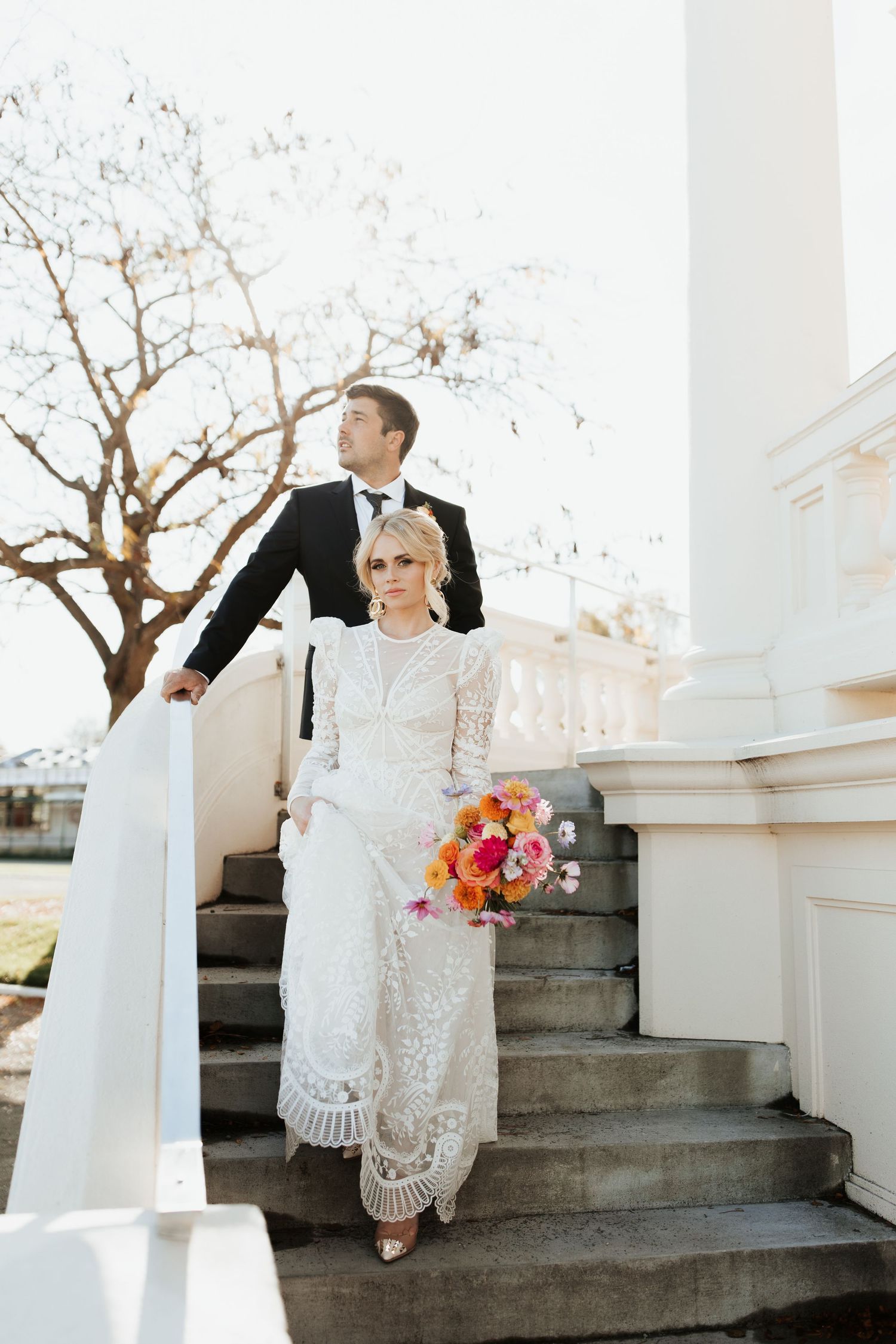Wedding couple stands on elegant stone steps, bride in lace dress holding vibrant coral and pink bouquet.