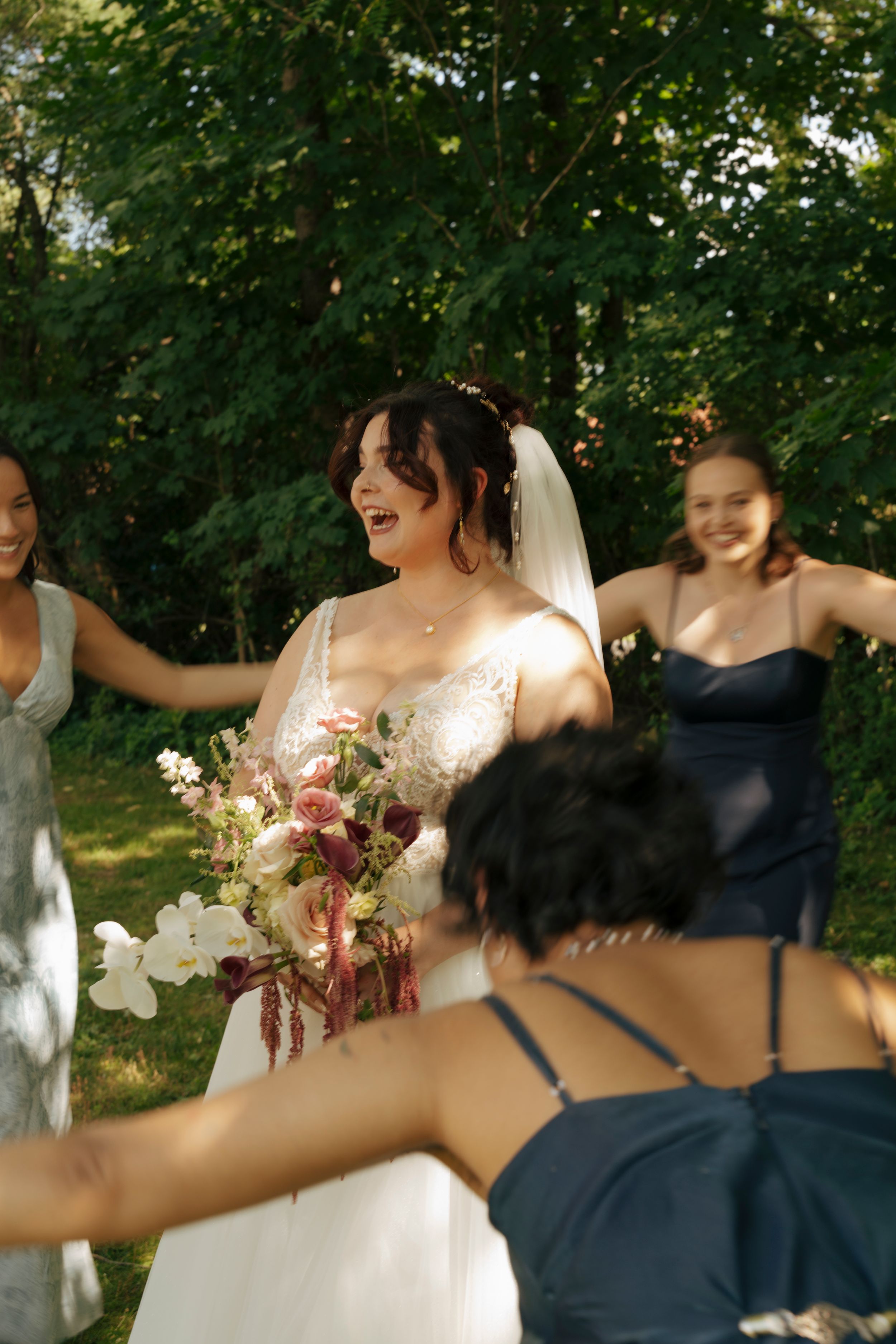 Bride in white lace gown holds floral bouquet while laughing with bridesmaids in navy dresses outdoors.