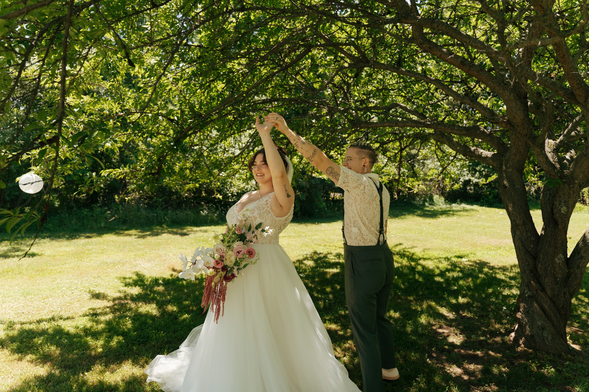 Bride and groom dancing outdoors under lush green trees, bride holding colorful bouquet in white gown.