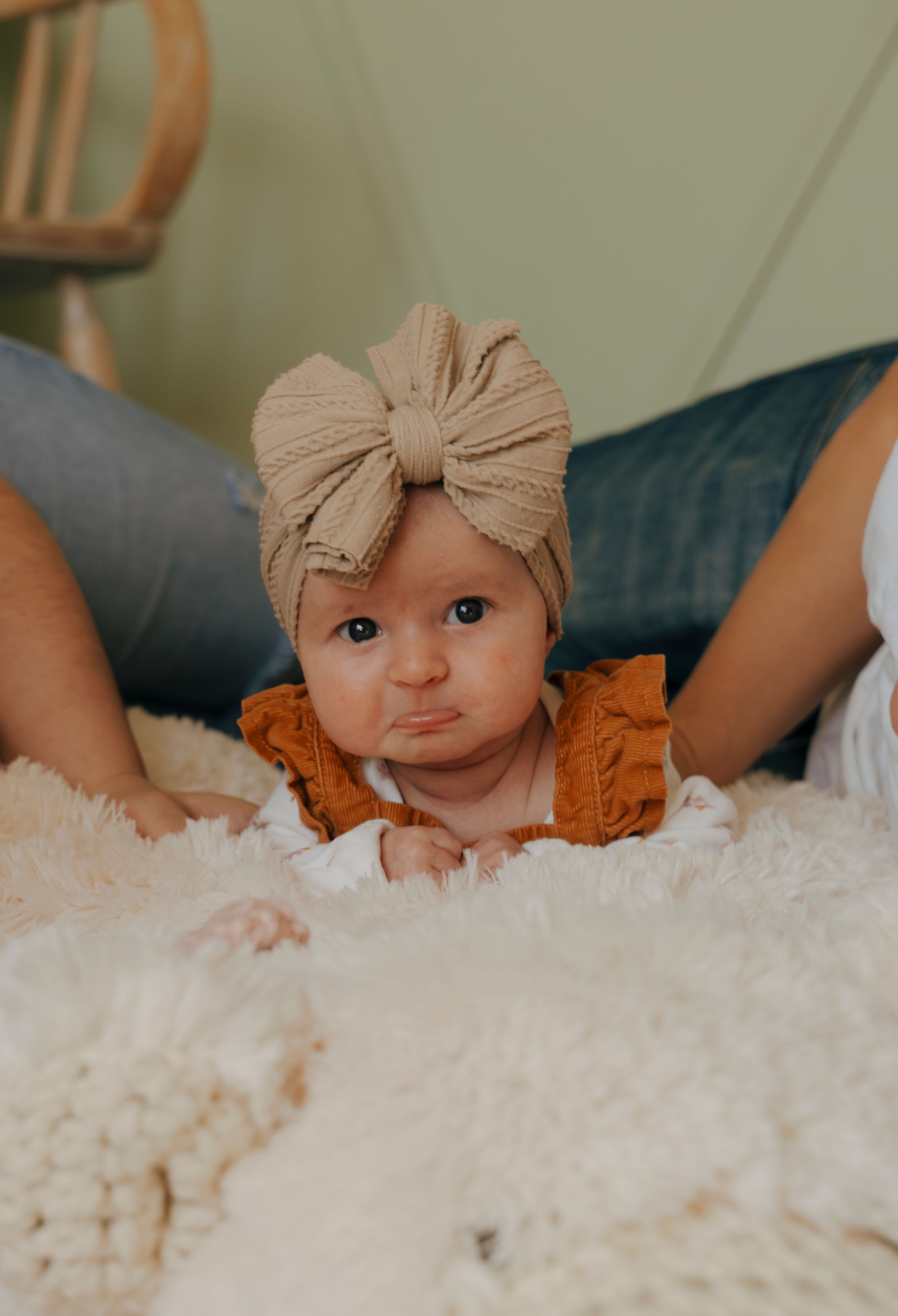A small baby wearing a beige bow headband and orange outfit lies on a fluffy white blanket.
