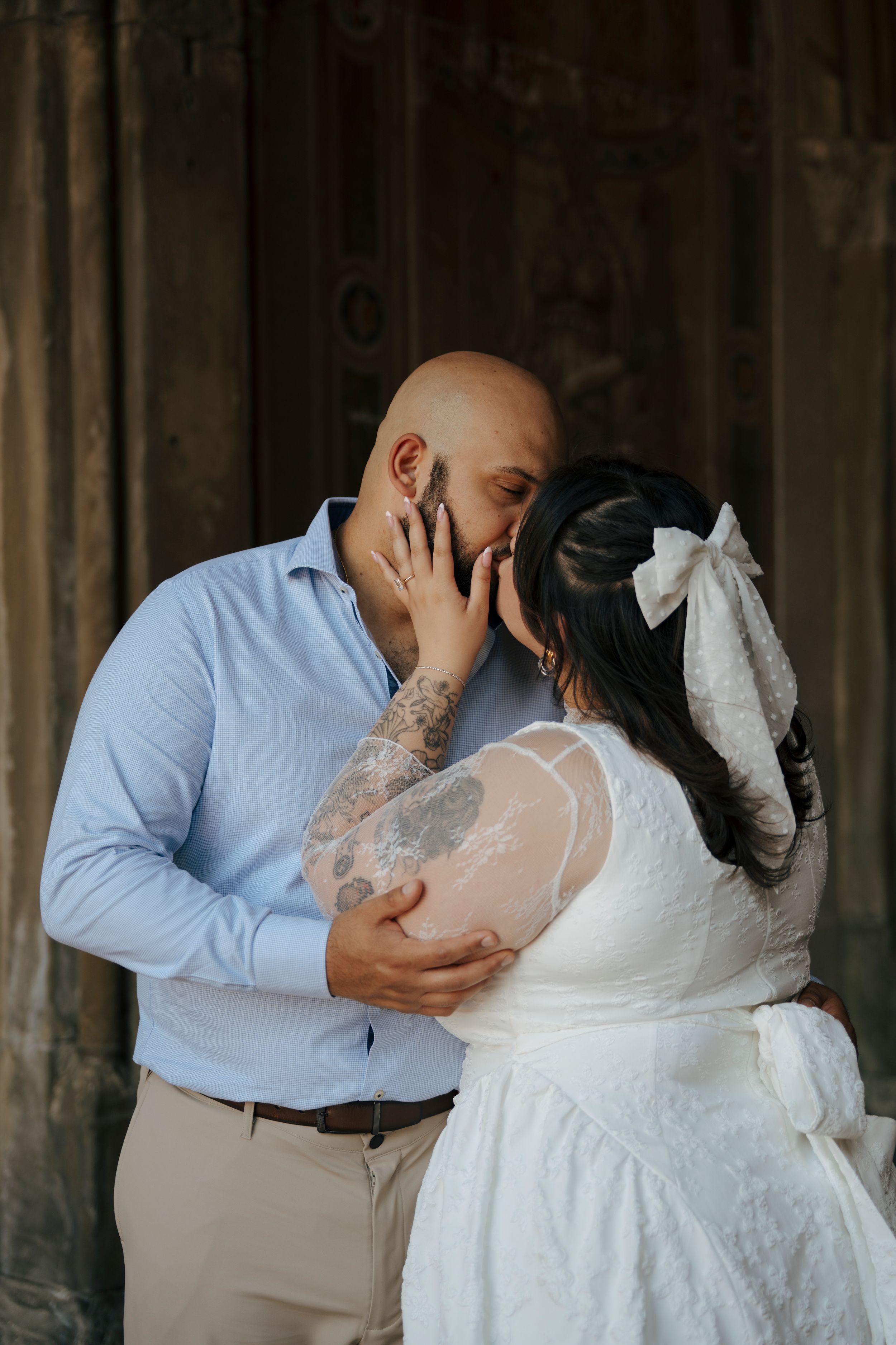 Couple sharing a romantic kiss, groom in blue shirt with tattoos embracing bride in white lace dress with bow.