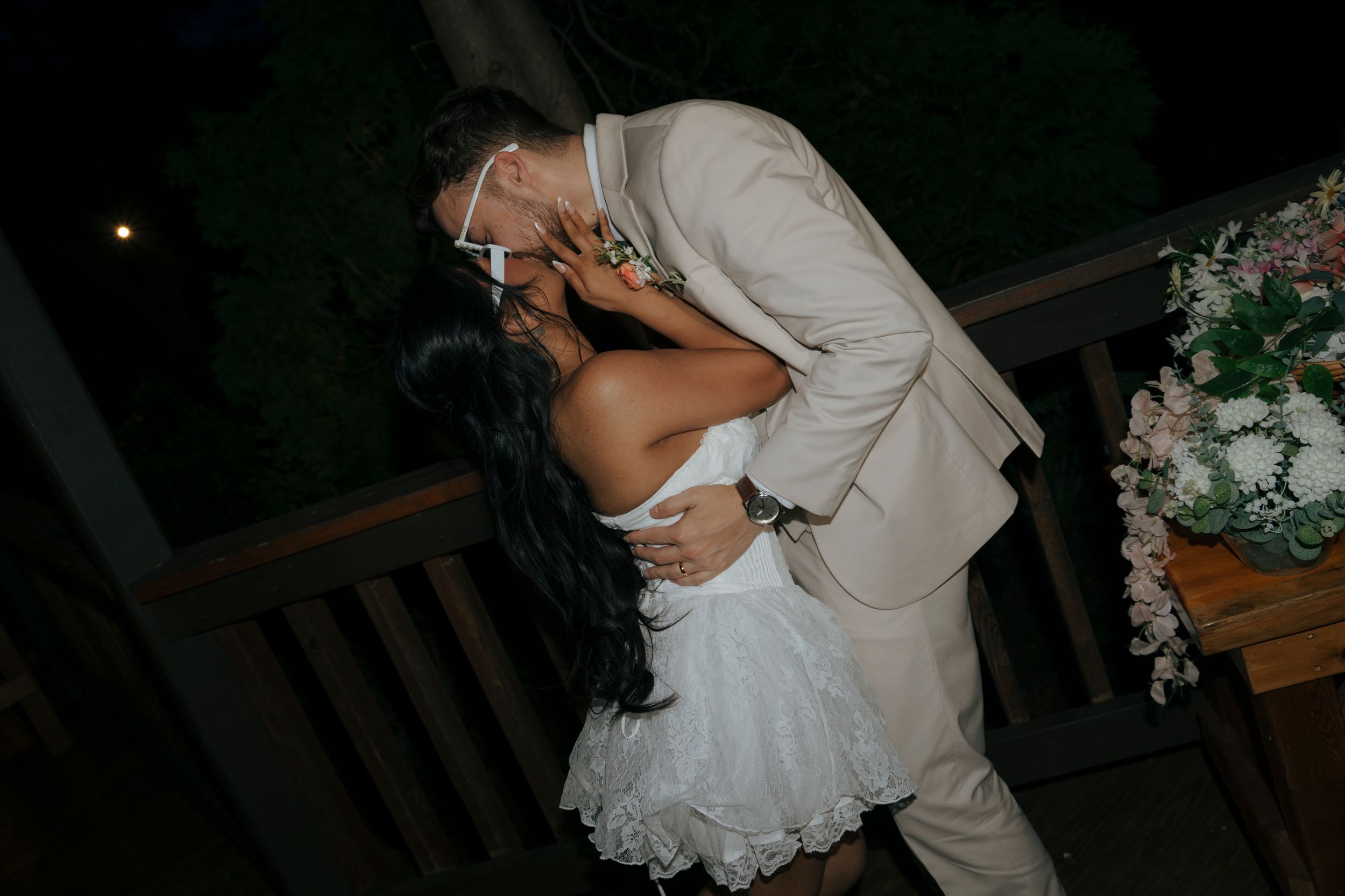 Bride and groom share a romantic dip kiss outdoors at night, bride in white dress, groom in tan suit.