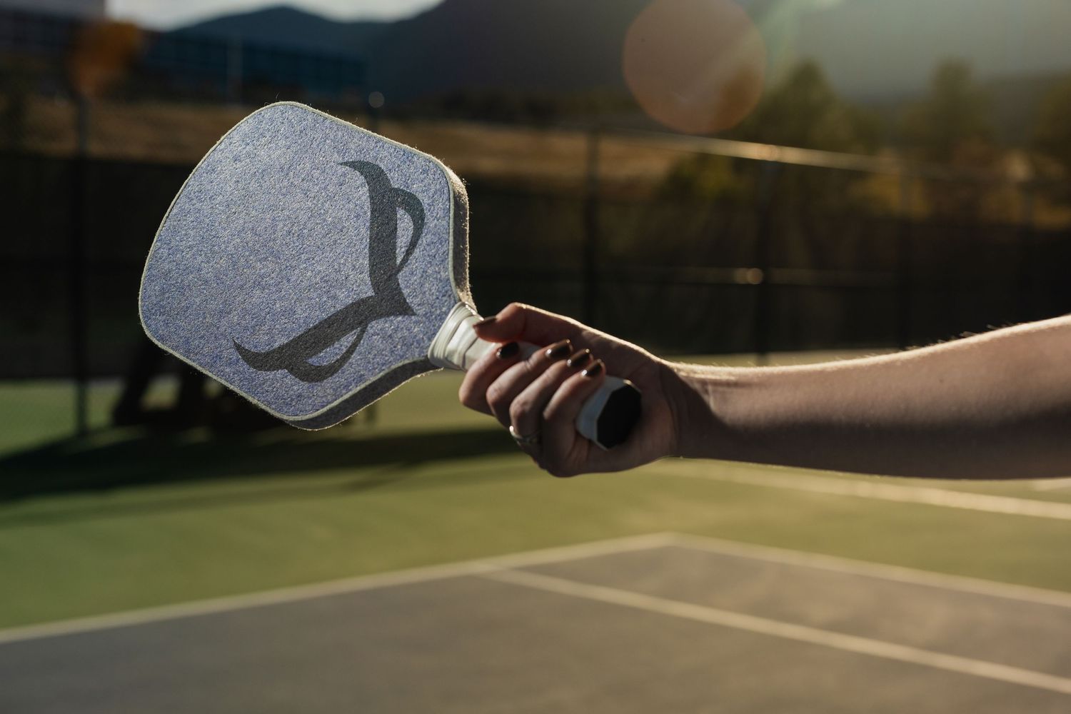 A hand holding a pickleball paddle on an outdoor court at sunset with lens flare.