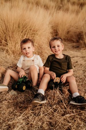 Children sit together in dry grass wearing casual clothes during a sunny outdoor photoshoot.