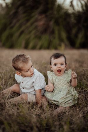 Two young siblings in light colored outfits playing together on dried grass at sunset.