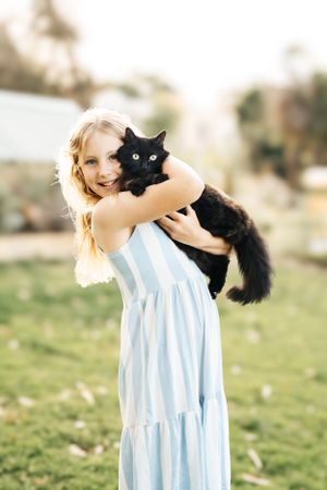 Young girl in light blue dress holding a black cat in an outdoor garden setting.
