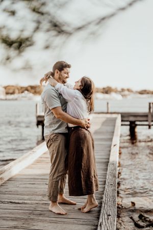Couple sitting close together on a wooden dock by the water at sunset.