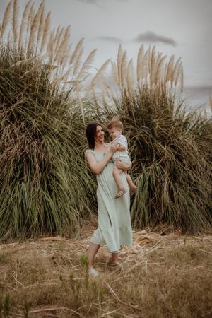 A woman in a mint dress holds a small child against a backdrop of towering pampas grass.
