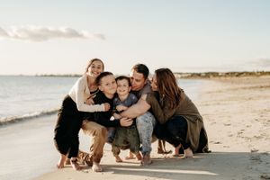Family members huddle together for a group hug on a sunny beach with ocean waves in the background.