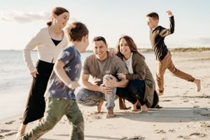 Family plays and laughs together on a sandy beach during golden hour with children running around.