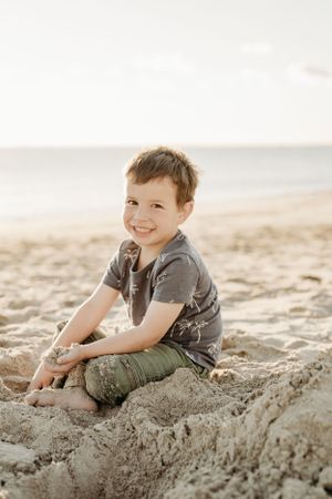 Young boy sits smiling on a sand dune at the beach during sunset.