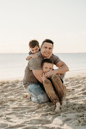 Family members share warm embrace while sitting together on sandy beach at sunset.