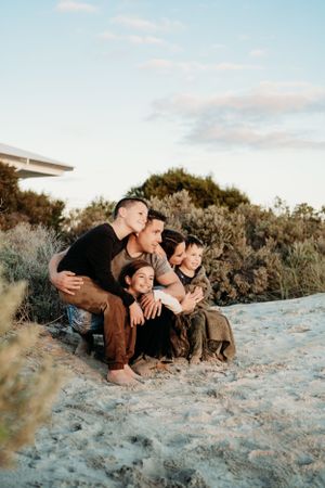 A group sits cuddled together on sandy ground against a sunset sky and coastal vegetation.