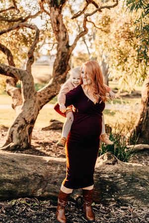 Candid maternity moment captured in golden sunlight under sprawling trees.
