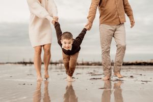 Playful family moment swinging child while walking barefoot on wet beach sand.