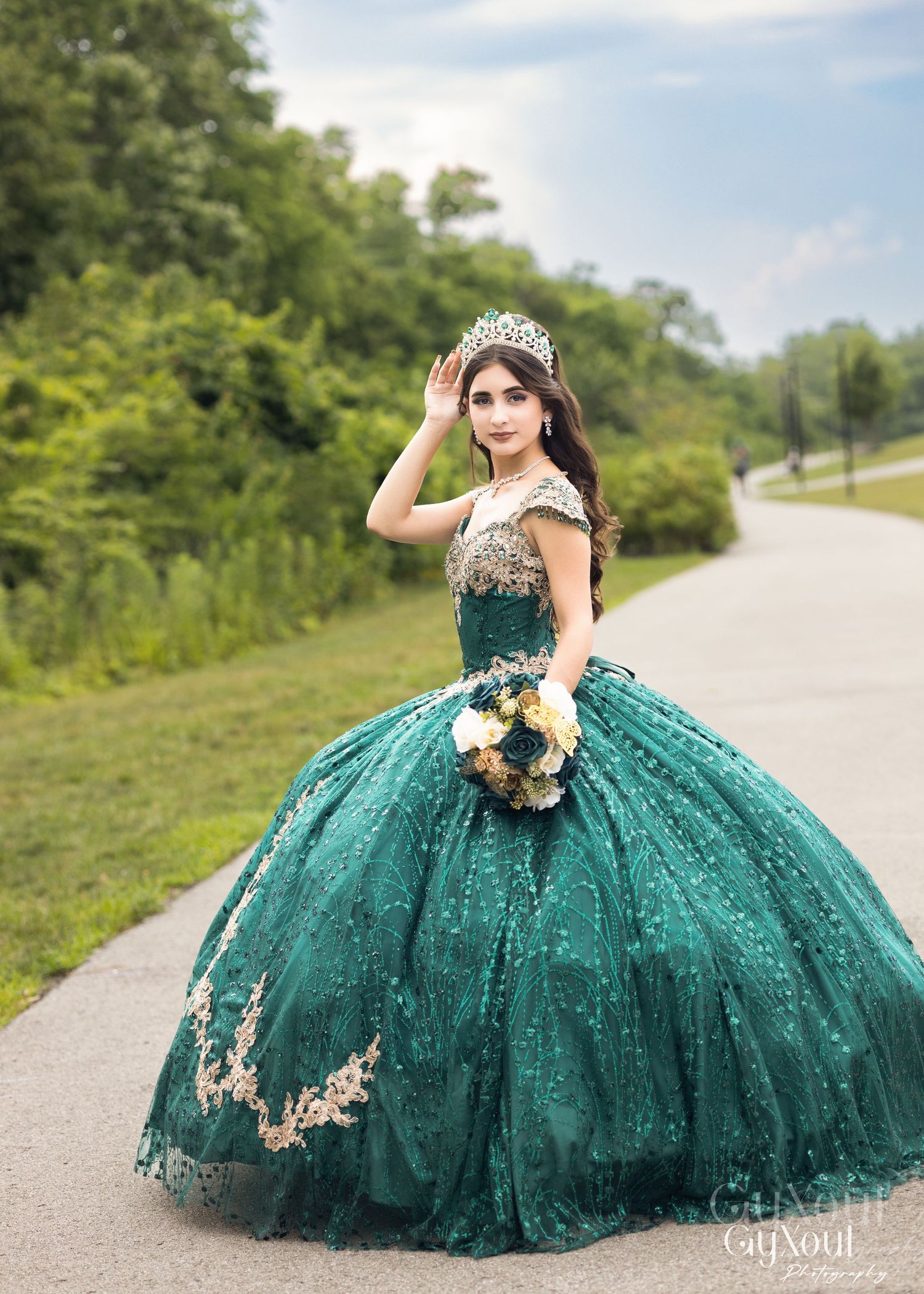 A feminine figure poses in an emerald green ball gown with gold embellishments and a bouquet in a scenic outdoor setting.